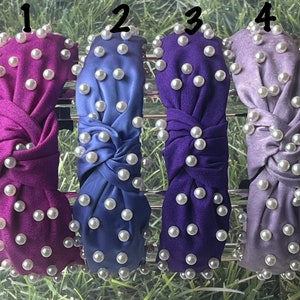May include: Four fabric headbands in shades of purple, blue, and lavender, each adorned with white faux pearls. The headbands have a twisted knot design and are displayed against a green and silver background.