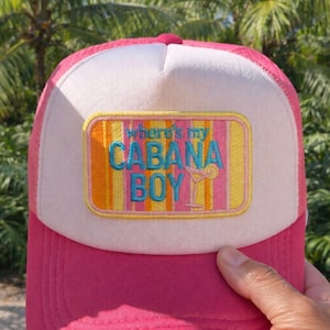 Puede incluir: Gorra de camionero rosa y blanca con la frase bordada "Where's my Cabana Boy" en la parte delantera. La gorra tiene una visera rosa y un panel frontal blanco con un parche rectangular colorido.
