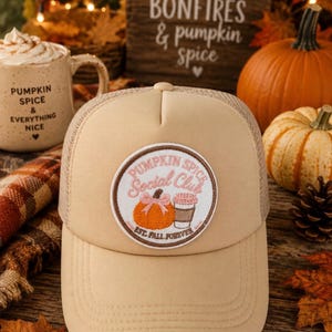 Puede incluir: Una gorra tipo trucker de color beige con un parche circular bordado en la parte frontal. El parche muestra el texto "PUMPKIN SPICE Social Club EST. FALL FOREVER" junto a una ilustración de una calabaza naranja con un lazo rosa y un café.