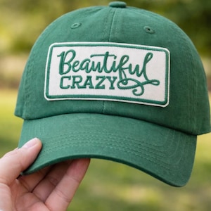 May include: A green baseball cap with a curved brim. The cap features a rectangular patch with the words "Beautiful Crazy" embroidered in white and green. The cap is made of a soft fabric and has a classic, casual style.