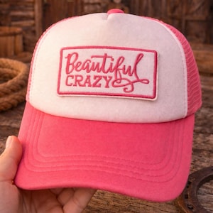 Może przedstawiać: Różowo-biała czapka typu trucker z zakrzywionym daszkiem. Czapka ma biały panel przedni z wyszytymi słowami "Beautiful Crazy" w kolorze różowym. Tylna część z siatki jest różowa, a czapka jest przeznaczona do noszenia na co dzień.