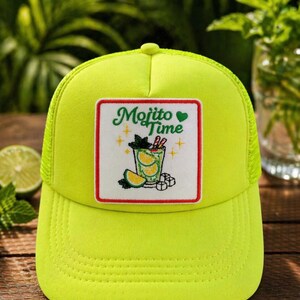 May include: A neon green trucker hat with a mesh back and a curved brim. The front features a square patch with the words "Mojito Time" and a graphic of a mojito cocktail with lime slices, mint, and ice cubes.