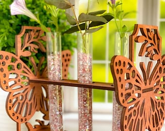 May include: Wooden butterfly-shaped flower vase with three glass test tubes filled with pink and white glitter. The vase is decorated with a floral design and has a brown finish.