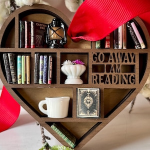 May include: A heart-shaped wooden bookshelf ornament with miniature books, a lantern, a teacup, and a small vase with flowers. The text "GO AWAY I AM READING" is cut out of the wood. A red ribbon is attached for hanging.