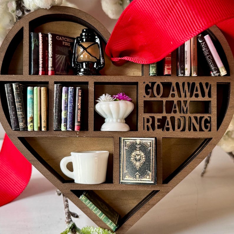 May include: A heart-shaped wooden bookshelf ornament with miniature books, a lantern, a teacup, and a small vase with flowers. The text "GO AWAY I AM READING" is cut out of the wood. A red ribbon is attached for hanging.