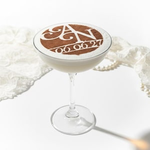 May include: A clear glass coupe cocktail filled with a creamy white liquid. The top foam features a brown cocoa powder stencil design displaying the letters AN and the date 06.06.21 in a circular frame, set on a thin, elegant glass stem.