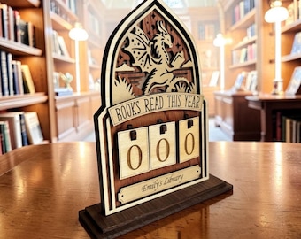 May include: Wooden book counter with a dragon design. The counter reads "BOOKS READ THIS YEAR" and displays "000". The base has the text "Emily's Library". The counter is made of light and dark brown wood.