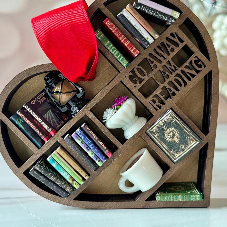 May include: A heart-shaped wooden shelf with compartments, filled with miniature books, a lantern, a small white vase with flowers, and a coffee cup. The words "GO AWAY I AM READING" are cut out of the wood. A red ribbon is attached to the top.