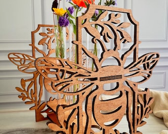May include: Wooden bee-shaped flower holders with intricate cut-out designs. The holders are made of light brown wood and feature a bee motif with wings and floral accents. Clear glass tubes hold colorful flowers, including orange poppies and purple lavender.