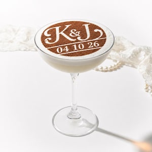 May include: A clear glass coupe cocktail glass filled with a creamy white beverage. The surface is topped with a brown cocoa powder stencil featuring the letters K&J and the date 04 10 26 in a decorative white font against the brown powder.