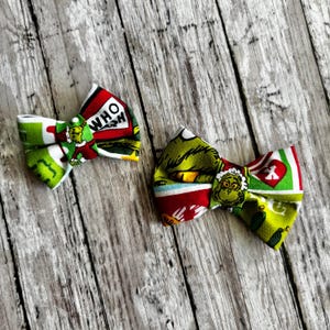 May include: Two fabric hair bows featuring a colorful print of a green character, red hearts, and the text WHO. The bows are constructed with folded fabric sections, showcasing a festive pattern with green, red, and white color blocks.