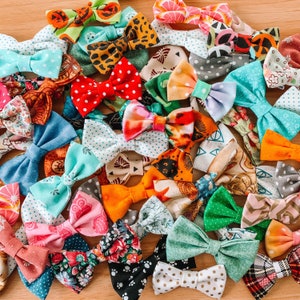 May include: A collection of colorful fabric bow ties in various patterns and colors, including polka dots, floral prints, and animal prints. The bow ties are arranged closely together, showcasing a variety of designs and textures. The color palette includes red, blue, green, and orange.