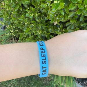 Peut inclure: Un bracelet en silicone bleu clair avec les mots "EAT SLEEP DIVE" imprimés en noir. Le bracelet est porté au poignet d'une personne, avec un fond de feuilles vertes.