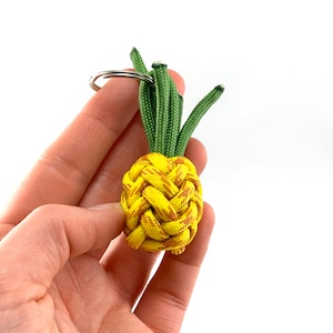 May include: A yellow and brown pineapple-shaped keyring with green leaves. The keyring is made of paracord and has a silver metal ring.