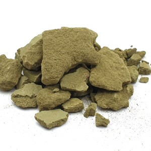 Pode incluir: Um monte de rochas verde-oliva de formato irregular sobre um fundo branco. As rochas t&ecirc;m superf&iacute;cies &aacute;speras e texturizadas e variam em tamanho. Fragmentos menores est&atilde;o espalhados na base da pilha.