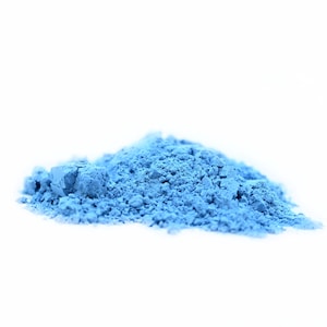 Puede incluir: Un pequeño montón de polvo azul claro sobre un fondo blanco. El polvo parece finamente molido y tiene una superficie ligeramente texturizada. El color es un azul pastel vibrante.
