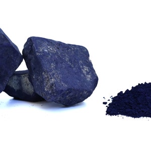 Op de afbeelding: Drie donkerblauwe indigo-verfstukken en een hoop indigopoeder. De verfstukken zijn ruw en onregelmatig van vorm. Het poeder is fijn en los.