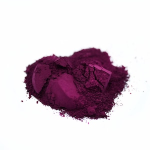 以下が含まれることがあります： 白い背景に、砕かれたマゼンタ色の粉末の山。粉末は細かく粉砕されているように見え、より大きな塊が見え、化粧品または顔料製品を示唆しています。色は深く、豊かな紫の色合いです。