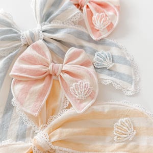 May include: Three fabric hair bows featuring pastel striped patterns in peach, light blue, and yellow. Each bow is accented with a small, white beaded seashell embellishment and finished with a delicate white lace trim along the edges.