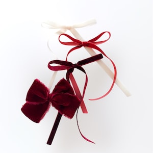 May include: An assortment of velvet bows in shades of red and cream against a white background. The bows vary in size and style, with some featuring long, flowing ribbons. The textures and colors create a visually appealing arrangement, suitable for gift wrapping or crafting.