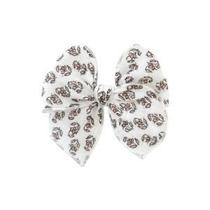 May include: A large, white fabric bow with a delicate floral pattern in shades of brown and tan. The bow is tied in the center and has a textured appearance, suitable for hair accessories or decorative purposes.