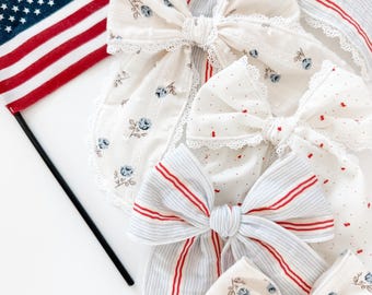 May include: A collection of five fabric hair bows featuring various patterns including blue floral prints, red and blue stripes, and red dots, all accented with delicate white lace trim, displayed alongside a small American flag on a black pole.