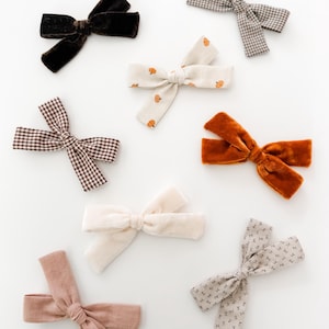 May include: A collection of eight hair bows in various textures and patterns, including dark brown velvet, beige with orange pumpkin prints, brown and white gingham, burnt orange velvet, solid cream, dusty pink, and light grey with small floral prints.