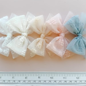 May include: A row of six pastel-colored tulle bows with a delicate floral pattern. The bows are in shades of white, cream, peach, and blue. A ruler is placed below the bows, with measurements in inches.