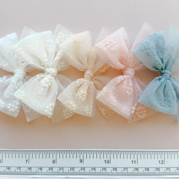 Puede incluir: Una fila de seis lazos de tul en colores pastel con un delicado estampado floral. Los lazos son en tonos blanco, crema, melocotón y azul. Una regla con medidas en centímetros se coloca debajo.