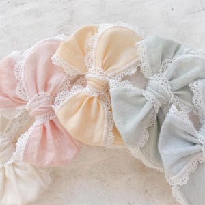 Puede incluir: Una colección de cuatro lazos de tela para el cabello en colores pastel suaves que incluyen rosa claro, amarillo pálido, verde salvia y blanco hueso. Cada lazo presenta un nudo central y está bordeado con un delicado encaje blanco superpuesto.