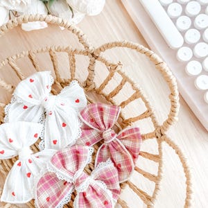 May include: A woven basket holds several fabric hair bows. Two white bows feature red heart patterns and lace trim. Two pink plaid bows with red heart accents and lace trim are also in the basket. A keyboard is visible in the background.