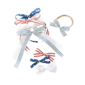 Puede incluir: Una colección de accesorios para el cabello con varios lazos en distintos estampados, incluyendo azul con estrellas blancas, rayas rojas y blancas, cuadros vichy azul claro y encaje blanco. El set incluye varias pinzas y una diadema.