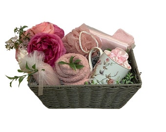 May include: A gray wicker basket filled with pink bath products, including a mug, towels, and a bath puff. The basket is decorated with pink flowers.