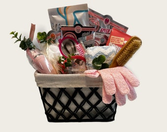 May include: A black and white woven basket filled with a variety of bath and body products, including a pink bath mitt, a wooden brush, a pink and white bath pouf, a pink and white face mask, a pink and white body scrub, and a pink and white body lotion.