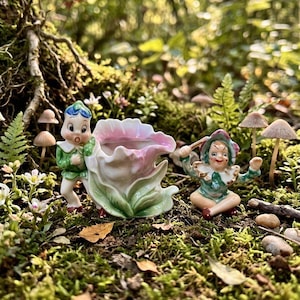 May include: A ceramic planter shaped like a large white and pink flower, accompanied by two small elf figurines. One elf stands beside the flower with a surprised expression, while the other sits on the ground holding a small stick-like object.