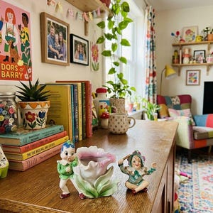 May include: A ceramic planter shaped like a pink and green tulip with a small elf figure attached to the side, positioned next to a small, seated elf figurine wearing a green and purple jester-style outfit on a wooden surface.