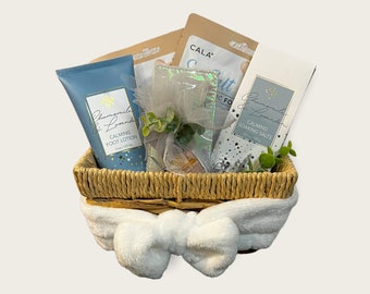 May include: A wicker basket filled with a blue and white foot lotion, a blue and white soaking salts package, a silver and white foot mask, and a white headband. The basket is decorated with green leaves and a white bow.