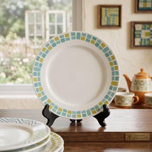 May include: A white ceramic dinner plate featuring a border of alternating light blue and lime green square tiles. The plate is displayed on a black stand. The text The Mosaic Collection - Artisan Dinnerware is visible at the top of the image.