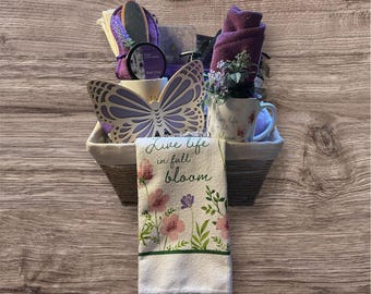 May include: A white wicker basket filled with a variety of bath and body products, including a purple towel, a wooden foot scrubber, a butterfly-shaped decoration, a mug with the text "Live life in full bloom", and a hand towel with a floral design and the text "Live life in full bloom".