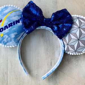 May include: Blue and white mouse ears with a blue sequined bow. The left ear features a blue and white cloud pattern with the text "SOARIN'" and a red and yellow logo. The right ear features a gray and white geometric pattern.