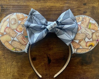 May include: A pair of gray and white mouse ears with a gray bow and a small bottle charm. The ears are decorated with a pattern of wine corks and the text "Wine Country".