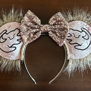May include: Pink velvet mouse ears with a brown fur trim and a gold glitter bow. The ears feature a black outline of Simba's paw print.