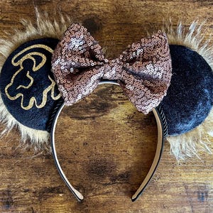 May include: Black velvet mouse ears with a gold lion silhouette and a rose gold sequin bow. The ears are trimmed with brown faux fur.