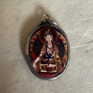 May include: Silver oval pendant with a colorful image of a deity in a seated position. The image is surrounded by a gold and red border.