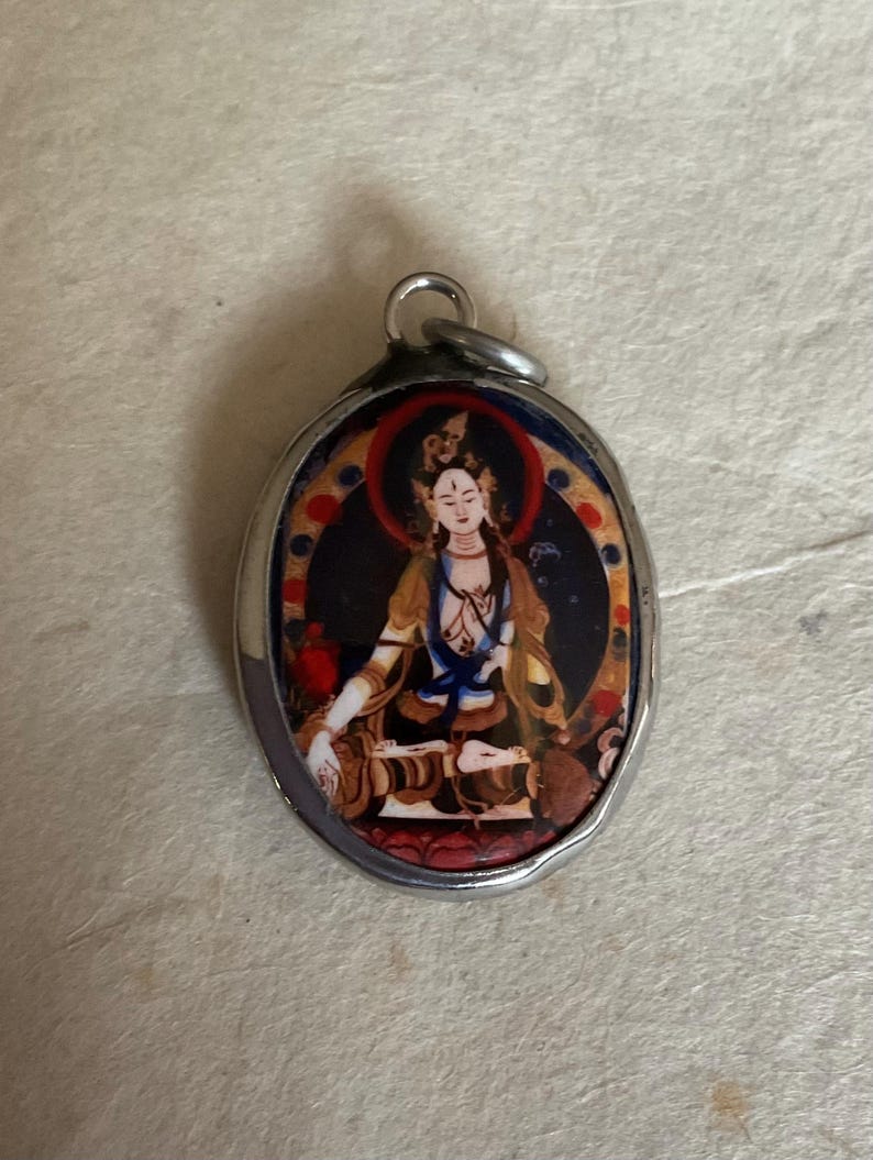 May include: Silver oval pendant with a colorful image of a deity in a seated position. The image is surrounded by a gold and red border.