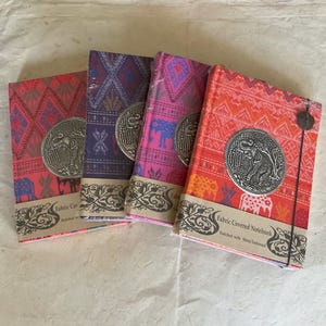 May include: Four fabric-covered notebooks with metal elephant embellishments. The notebooks have different colorful patterns and are labeled "Fabric Covered Notebook Embellished with Metal Embellishment".