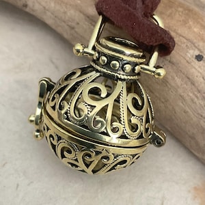 May include: A gold-colored, filigree pendant with an ornate, openwork design. The spherical pendant is suspended from a brown suede cord. The pendant has a hinged opening and a decorative top with a loop for the cord.