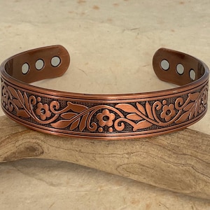 May include: A copper cuff bracelet featuring an embossed floral and vine pattern with a dark textured background. The interior of the open-ended band displays six small circular silver-toned magnets embedded near the ends of the bracelet.