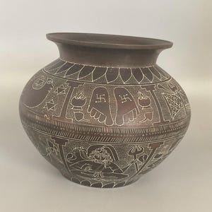 May include: A dark brown rounded pot featuring intricate white etched designs. The surface displays detailed engravings of human figures, a trident, footprints with swastika symbols, and various geometric patterns arranged in horizontal bands.