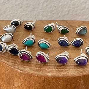 May include: A collection of silver stud earrings with various gemstone inlays. The earrings feature teardrop-shaped stones in colors like pink, white, black, green, blue, and purple. The earrings are displayed on a wooden surface.